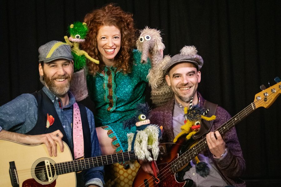 A group photo of 3 people with instruments and small monster puppets