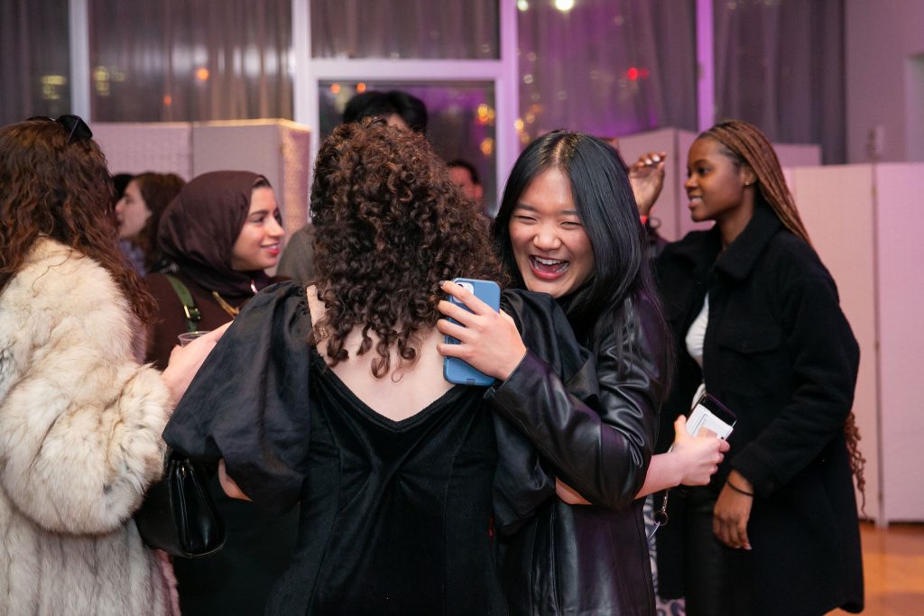 An Asian woman in all black smiles and hugs a woman of color in a black dress behind them three women talk to each other