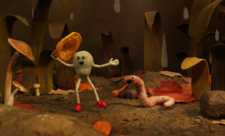 A claymation scene shows a round, green character with stick limbs and red shoes holding a yellow object, smiling at a happy pink worm in a whimsical forest filled with mushrooms and large leaves.