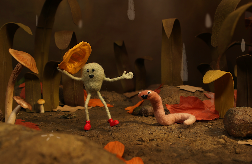A claymation scene shows a round, green character with stick limbs and red shoes holding a yellow object, smiling at a happy pink worm in a whimsical forest filled with mushrooms and large leaves.