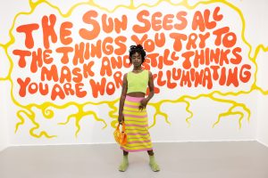 Dark skinned person in green and pink skirt and shirt outfit standing in front of orange and yellow text-based mural