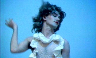 A video still showing a fair-skinned person in a white dress and red lipstick in motion.