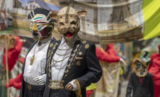 Two masked sculptures in matching black suits with gold adornments in front of a large banner