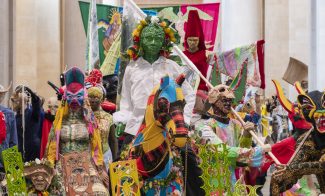 A procession of sculpted figures with colorful clothing, flags, and masks.