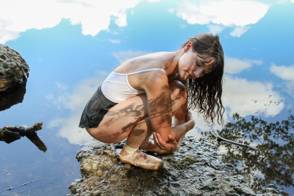 An image of a young woman in ballet clothing crouching at the edge of a body of water with the sky reflecting on the surface.