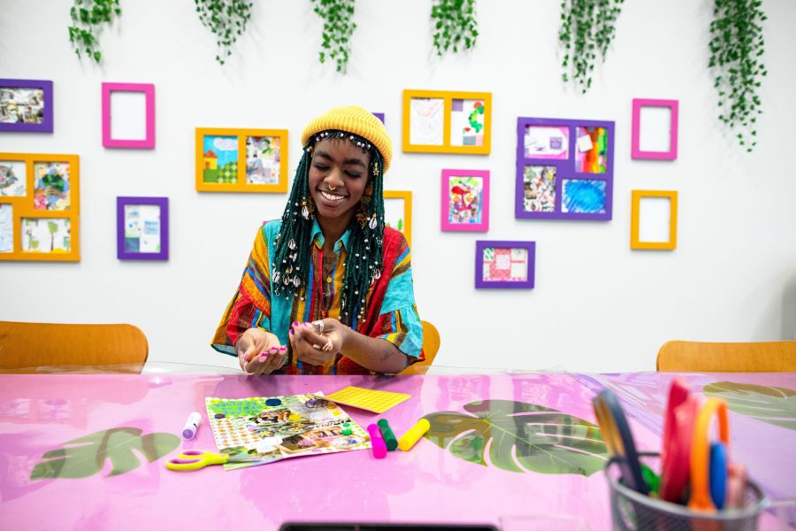 A person with long beaded braids and a yellow hat smiles as they create a 2-D artwork