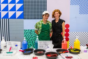 a man in white hat, a green apron, and underneath it, an abstract green shirt stands behind a table with art supplies. beside him, a woman with red hair in a black polka dot top and black apron smiles 