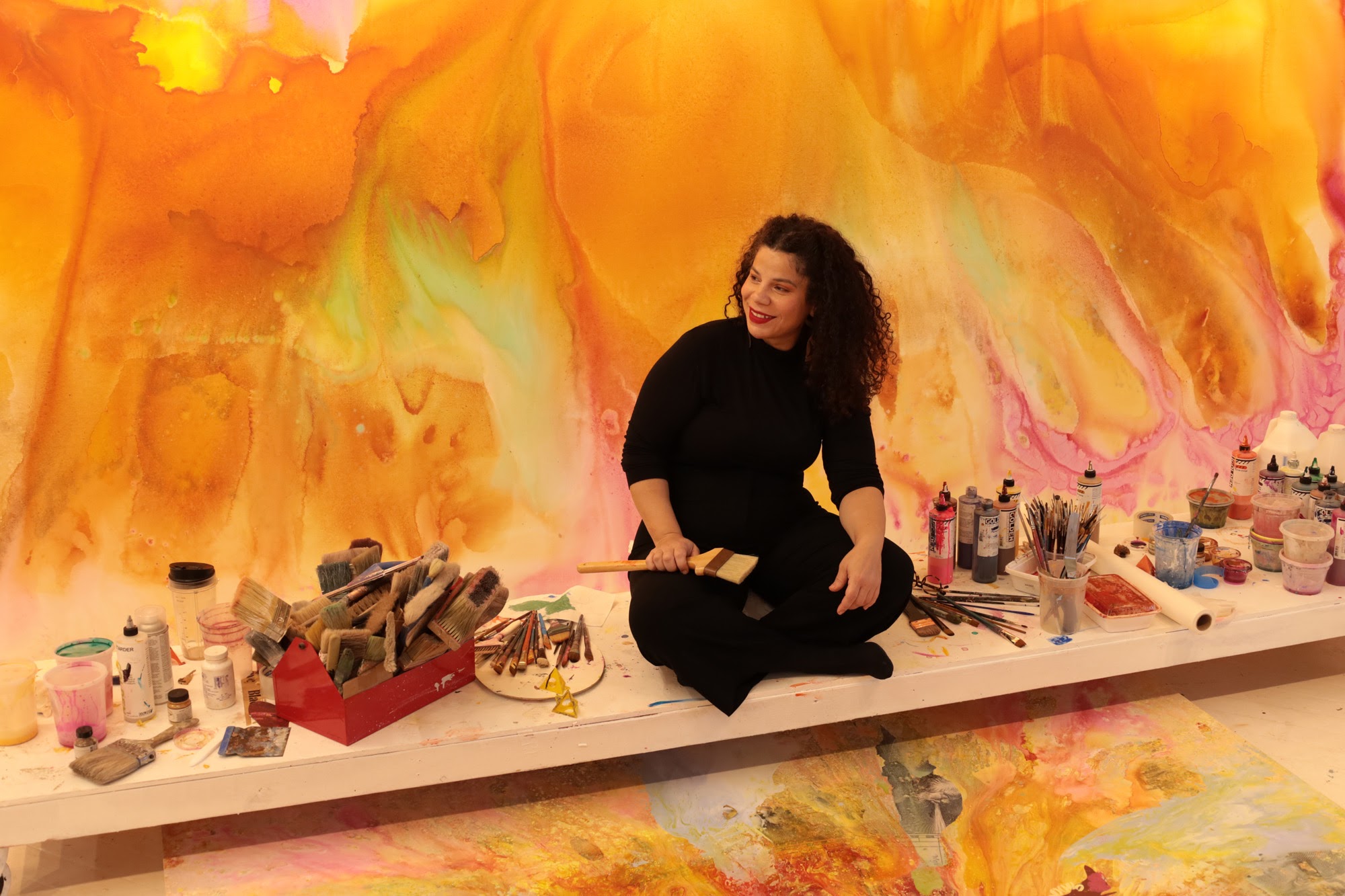 An artist with shoulder-length curly hair sitting in the studio painting a large orange painting
