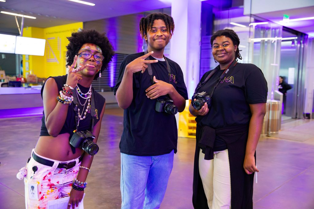 Three teens stand inside the ICA's lobby, each wearing black shirts and holding cameras. They smile for the photo, with one flashing a peace sign. Bright lights and modern architecture hint at a vibrant teen night in the background.