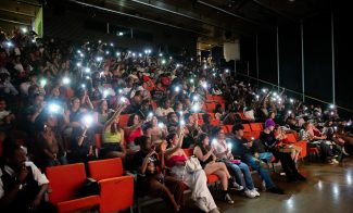 People sitting in orange theater seats in a dark theater watching a performance and shining their flashlights on the performers.