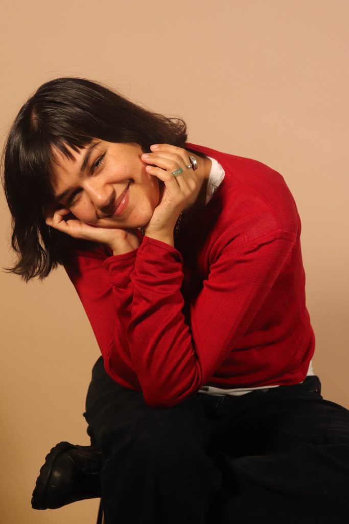 A person with short dark hair, wearing a red long-sleeve shirt and black pants, smiles while resting their face in their hands, sitting against a plain beige background.