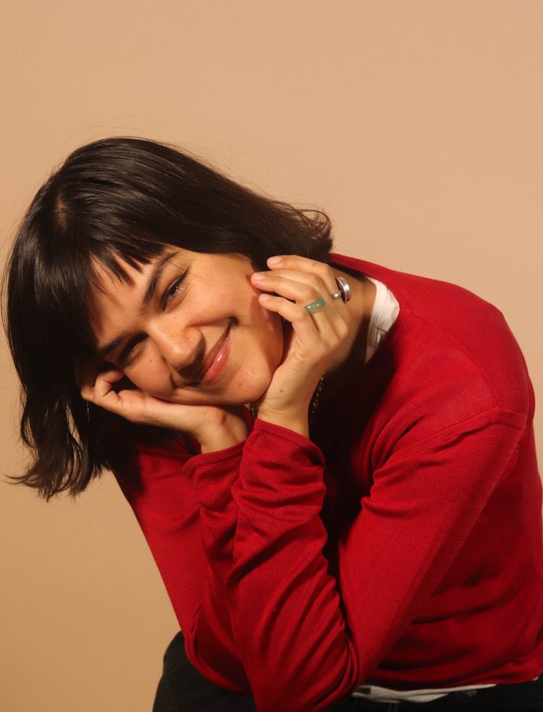A person with short dark hair, wearing a red long-sleeve shirt and black pants, smiles while resting their face in their hands, sitting against a plain beige background.