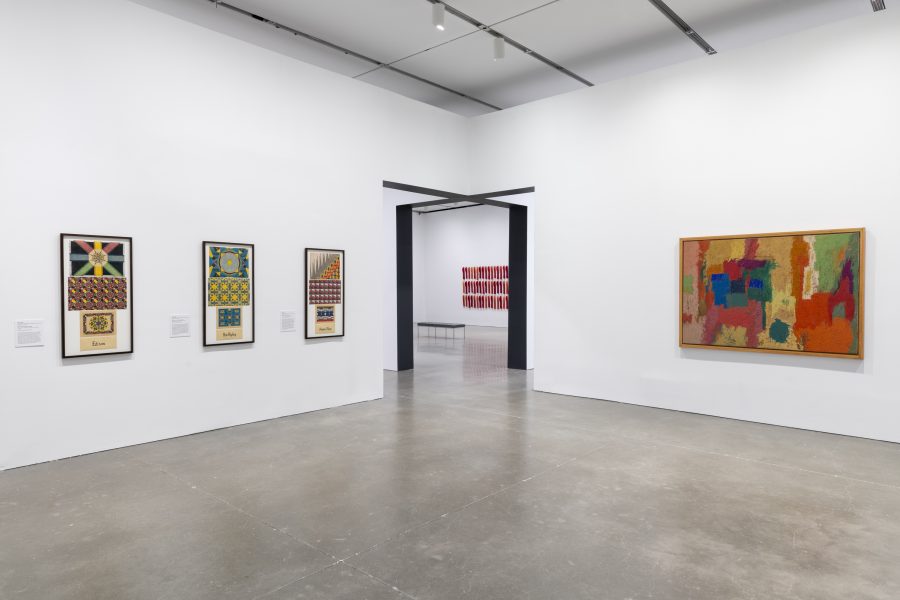 A gallery with white walls displays colorful abstract paintings and patterned drawings in simple frames; a large doorway offers a glimpse of more art in the next gallery