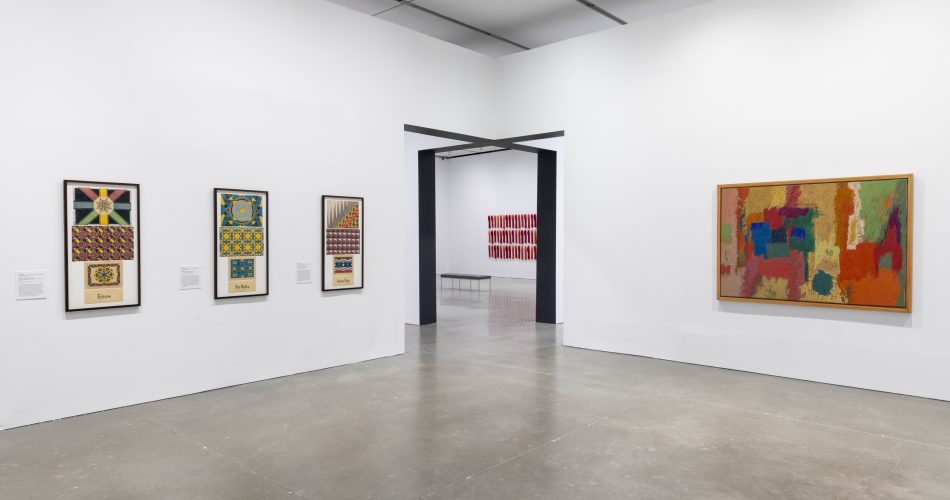A gallery with white walls displays colorful abstract paintings and patterned drawings in simple frames; a large doorway offers a glimpse of more art in the next gallery