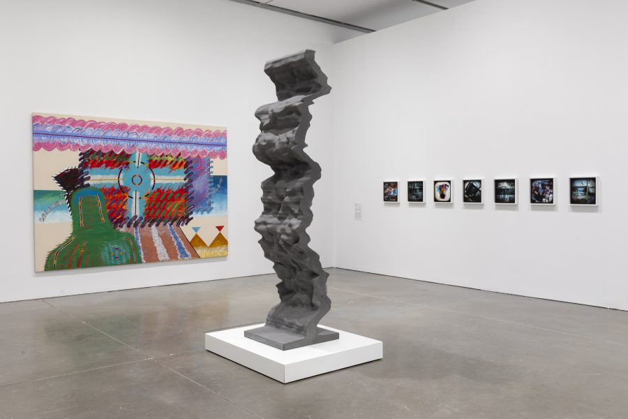 A tall, abstract gray sculpture on a white platform, a colorful abstract painting on the left wall, and a row of small framed photographs on the right wall