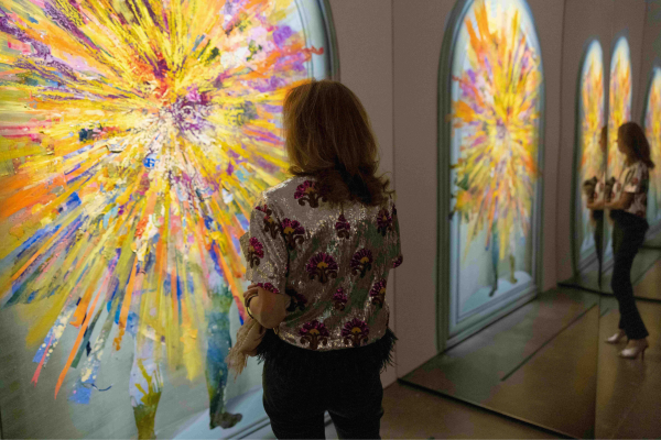 a woman in a silver sequin shirt stands in front wall length, painting of a man bursting with colors.