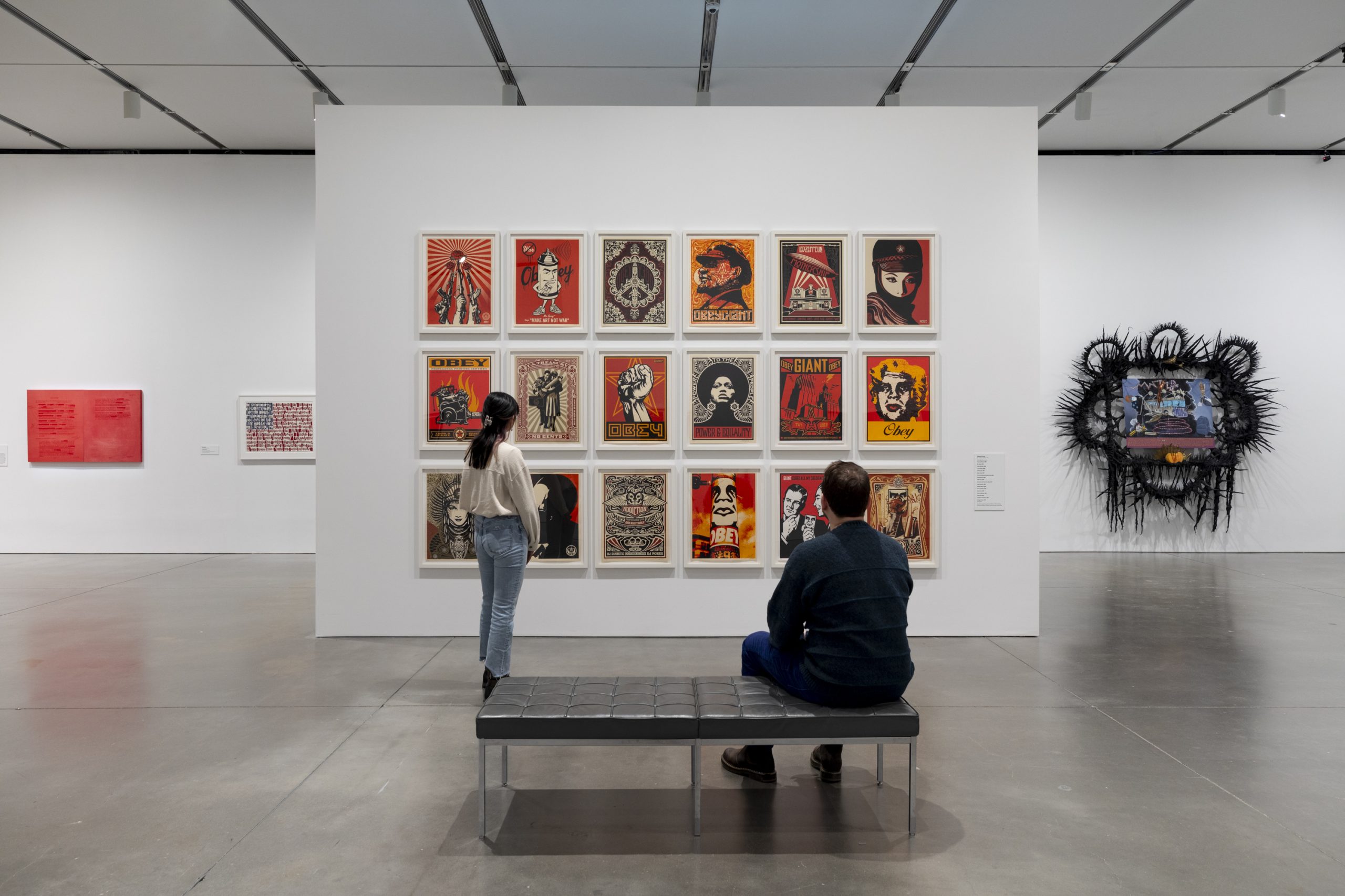 a man sits on a black leather bench and a woman stands looking at a work of multiple prints on a white gallery wall