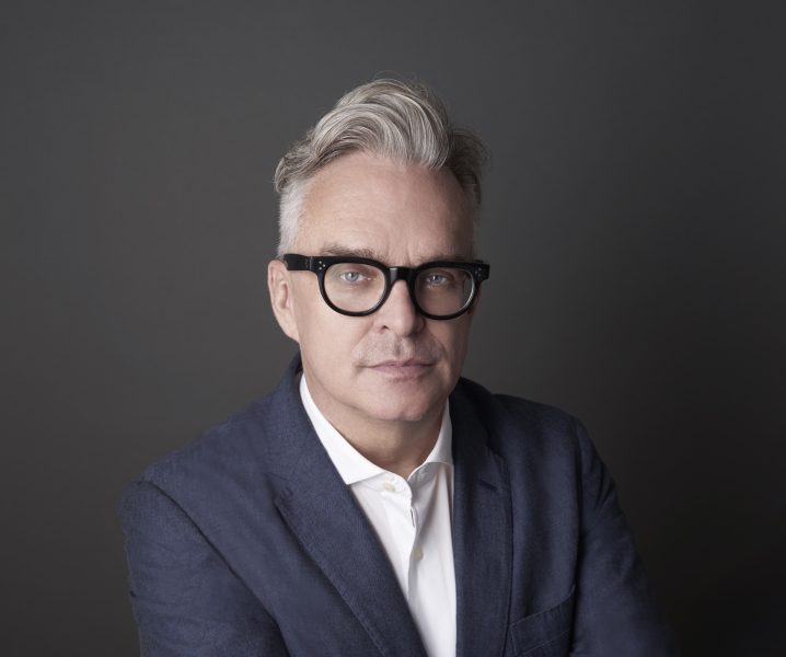 a man with gray hair in black rimmed glasses and a blue sports coat smirks for a photo against a gray background