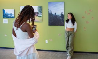 A dark-skinned person wearing a white top, skirt, and sweater uses their phone to take a photo of a medium-skinned person wearing a white top and green pants standing in front of works of art on a gallery wall.