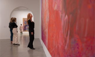 a woman in a black outfit looks up at a red and pink painting in a gallery space. behind her two women are in conversation