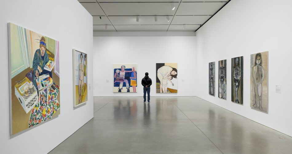 A person stands in a gallery, viewing various large paintings on white walls. The pieces include abstract and figurative works, displayed in a spacious room with a polished concrete floor and a ceiling with spotlights.