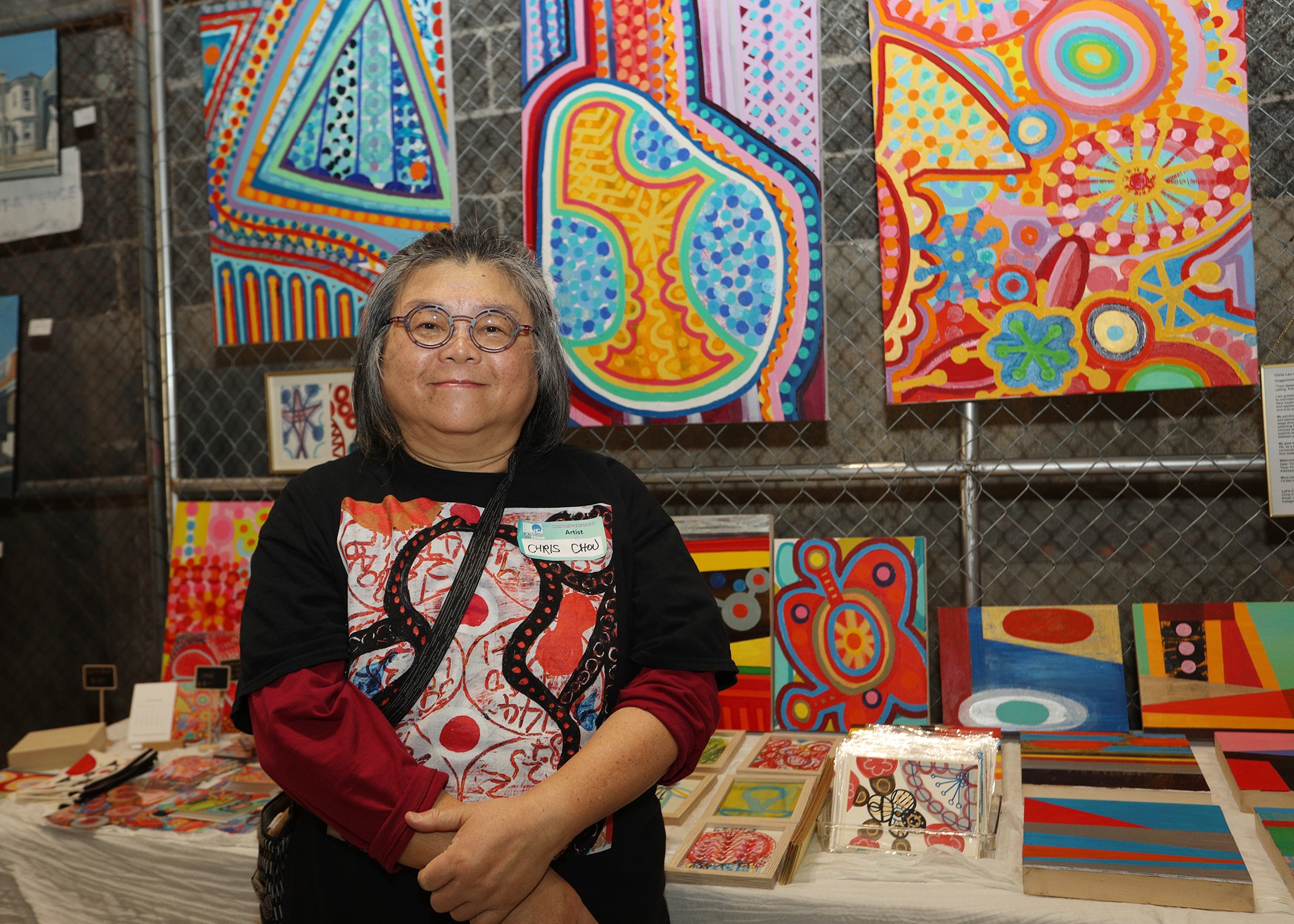A person with long gray hair and glasses stands in front of a display of colorful abstract paintings at an art show, smiling and wearing a black and red shirt. Artworks are arranged on the wall and table.