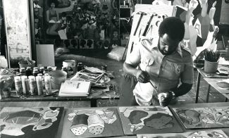 A man paints at a table covered with canvases and art supplies in a busy studio. Colorful abstract and figurative artworks, reflecting the vibrant spirit of AAMARP, are displayed on the wall behind him.
