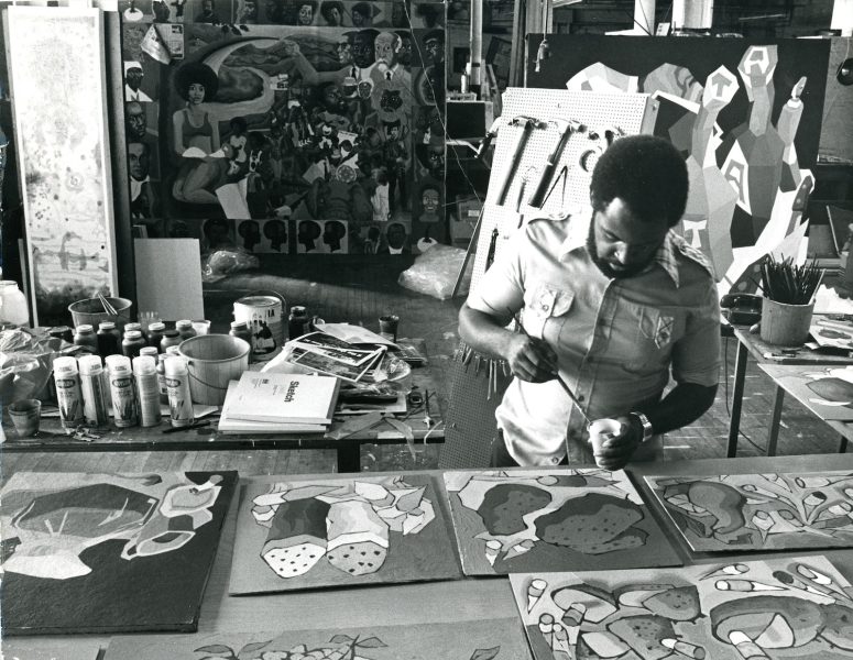 A man paints at a table covered with canvases and art supplies in a busy studio. Colorful abstract and figurative artworks, reflecting the vibrant spirit of AAMARP, are displayed on the wall behind him.