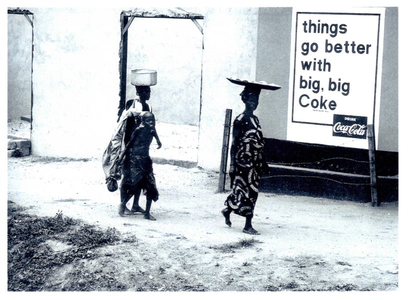 Two people walk by a wall with a Coca-Cola advertisement that reads, things go better with big, big Coke. One carries a bowl on their head, the other carries a bundle on their back.