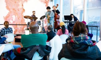 A jazz quartet, reminiscent of the Gregory Groover Jr Trio, performs in a modern venue. The musicians play drums, saxophone, double bass, and keyboard. An audience in casual attire listens intently as light streams through large windows onto coral-colored wall art.