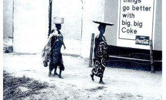 Two women and a child walk by a building in a rural area, balancing items on their heads. A large Coca-Cola sign reads, things go better with big, big Coke—an image reminiscent of works featured at AAMARP ICA Boston.