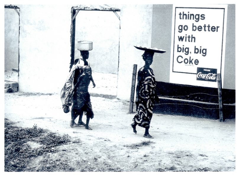 Two women and a child walk by a building in a rural area, balancing items on their heads. A large Coca-Cola sign reads, things go better with big, big Coke—an image reminiscent of works featured at AAMARP ICA Boston.