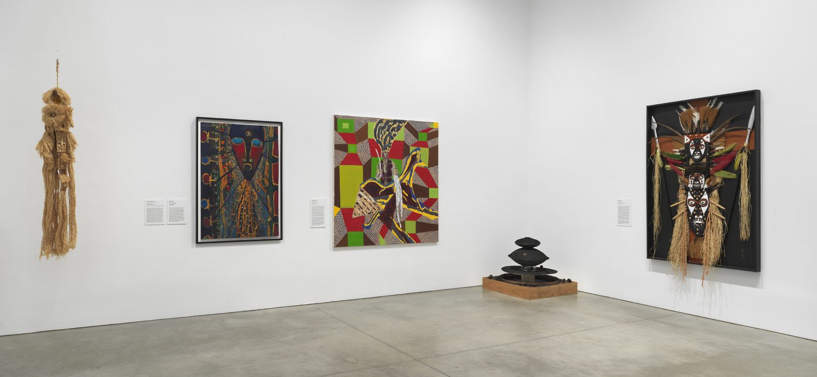 A gallery with white walls displays five artworks, including abstract paintings, a geometric sculpture on the floor, and a straw wall hanging