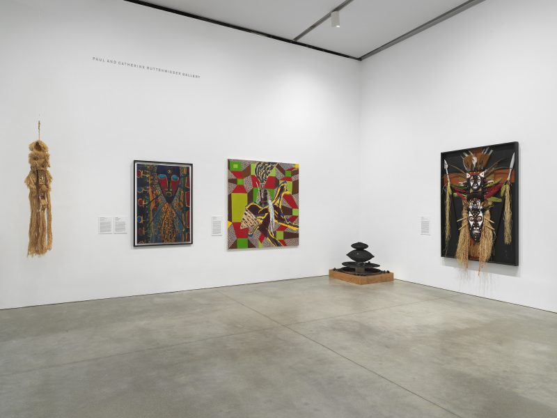 A gallery with white walls displays five artworks, including abstract paintings, a geometric sculpture on the floor, and a straw wall hanging