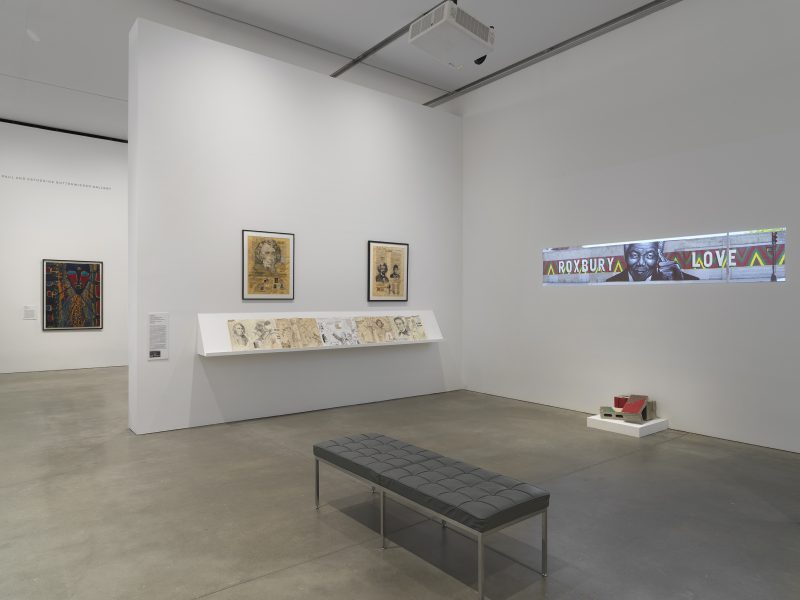 A gallery features framed artworks, sketches on a ledge, a digital projection of a mural depicting Nelson Mandela with text “Roxbury Love,” a small sculpture, and a black bench on a polished concrete floor