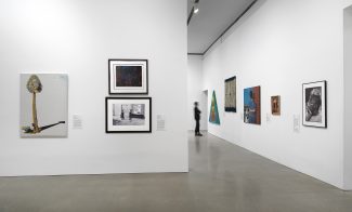 A gallery with white walls displays various paintings and framed artworks. A person in dark clothing walks in the background near a hallway
