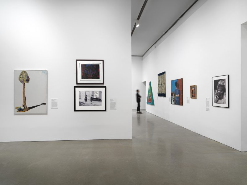 A gallery with white walls displays various paintings and framed artworks. A person in dark clothing walks in the background near a hallway