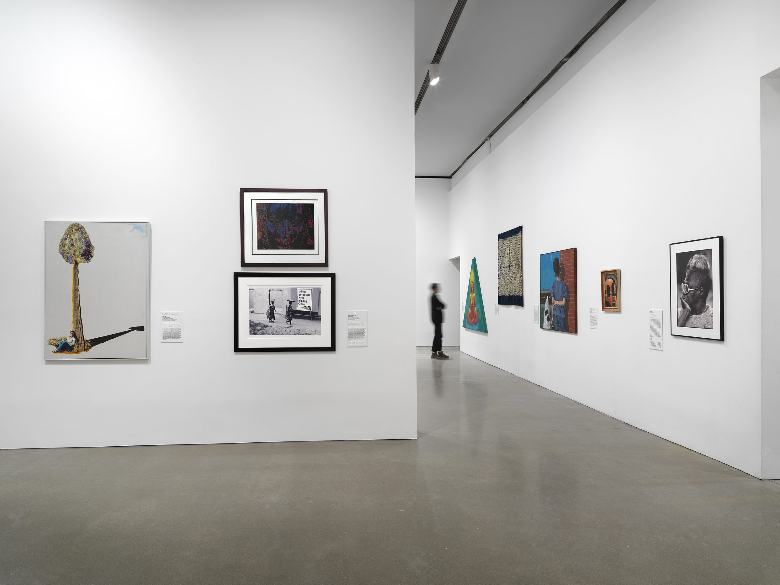 A gallery with white walls displays various paintings and framed artworks. A person in dark clothing walks in the background near a hallway