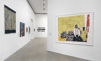 A gallery with white walls displays various artworks, including a large painting of two people on a patterned couch. On the far wall, black text reads “Say It Loud: AAMARP 1977 to Now.”.