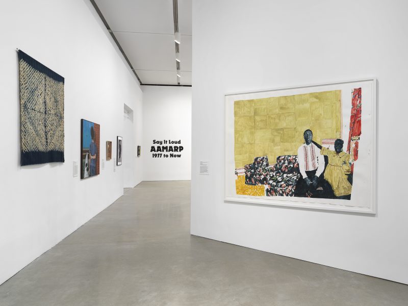A gallery with white walls displays various artworks, including a large painting of two people on a patterned couch. On the far wall, black text reads “Say It Loud: AAMARP 1977 to Now.”.