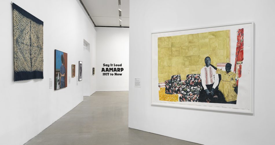 A gallery with white walls displays various artworks, including a large painting of two people on a patterned couch. On the far wall, black text reads “Say It Loud: AAMARP 1977 to Now.”.