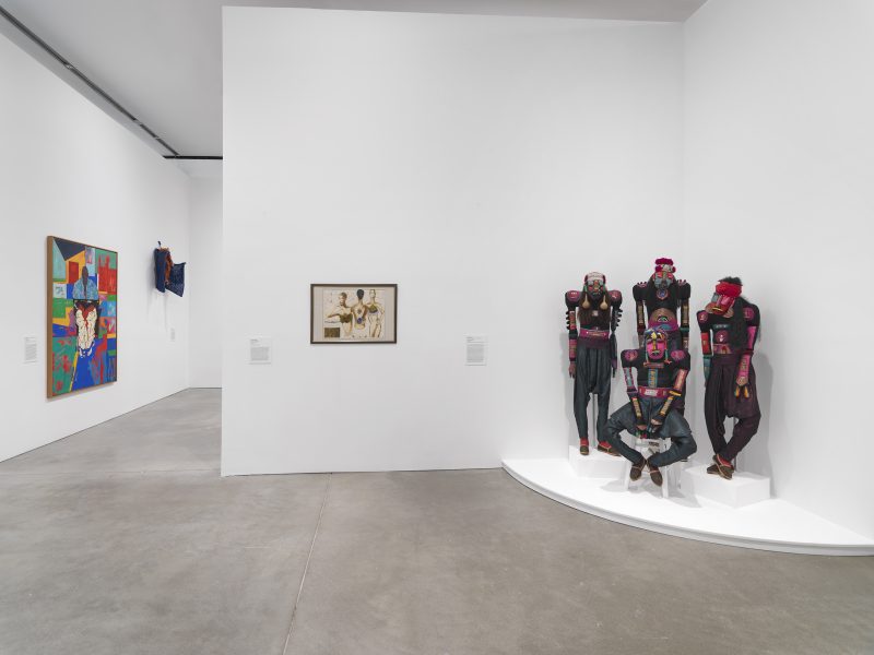 A gallery features colorful paintings on white walls and a sculptural group of four life-sized figures with face paint and vibrant costumes posed on a white platform in the corner.