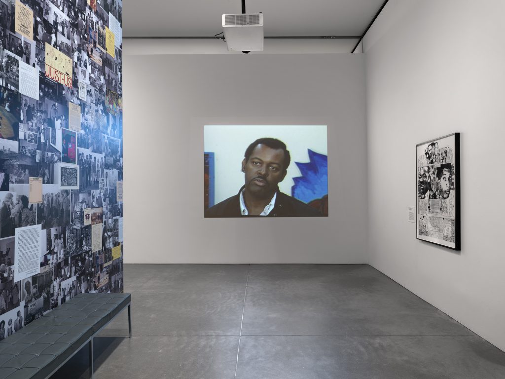 An art gallery features a video projected on a white wall, showing a man in a black jacket. The left wall displays black-and-white photos and documents, including works from AAMARP. A bench sits below; another artwork hangs right.