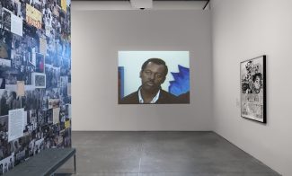 An art gallery features a video projected on a white wall, showing a man in a black jacket. The left wall displays black-and-white photos and documents, including works from AAMARP. A bench sits below; another artwork hangs right.
