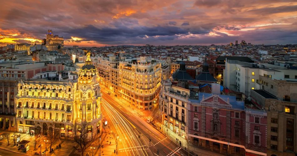 A stunning cityscape of Madrid at sunset, with vibrant lights illuminating the streets and historic buildings. The sky is filled with dramatic clouds, adding to the picturesque view of the bustling city below.