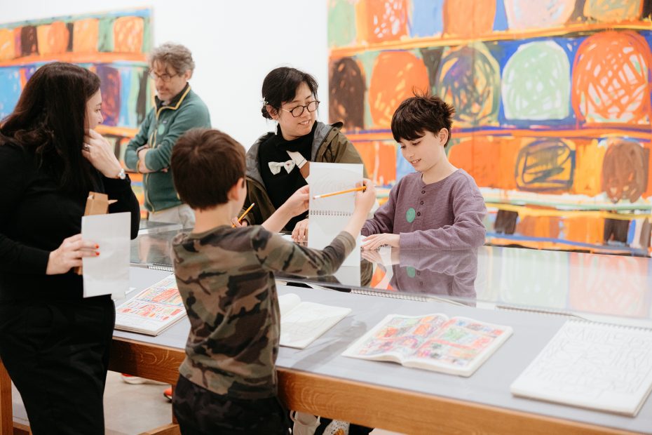 A group of people, including children and adults, gather around a display table with open sketchbooks as colorful abstract art hangs on the wall behind them.