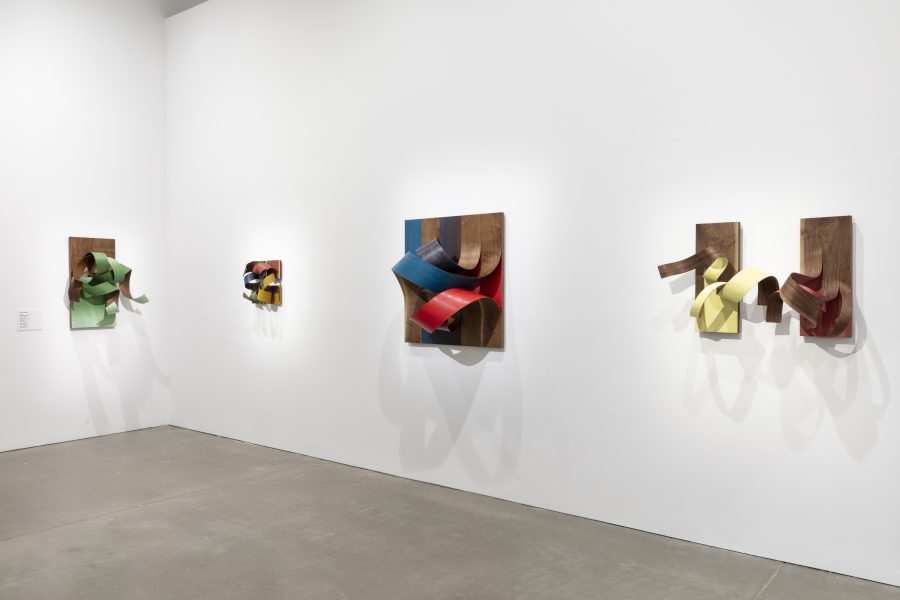 Art gallery with white walls displaying four abstract wooden wall sculptures. Each features curved, ribbon-like forms in bright colors on wood panels, casting dynamic shadows