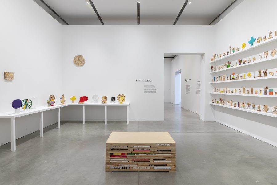 A gallery with white walls displays colorful small sculptures on tables and shelves. A low wooden bench with books stacked in its base stands in the center of the polished concrete floor.