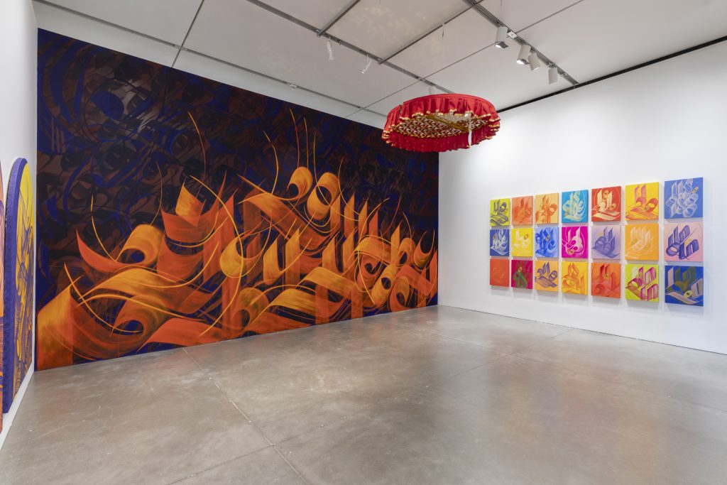 A gallery featuring a vibrant abstract orange and purple mural, colorful square paintings arranged in a grid, and a red circular ceiling installation