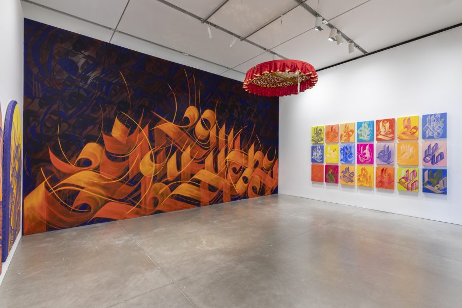 A gallery featuring a vibrant abstract orange and purple mural, colorful square paintings arranged in a grid, and a red circular ceiling installation